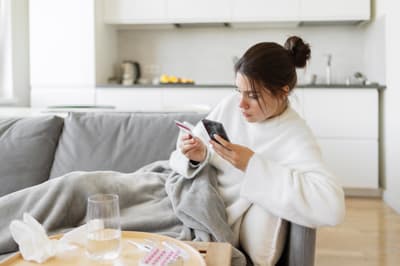 Erkältete Frau auf dem Sofa bucht mit Smartphone und Versichertenkarte einen Arzttermin