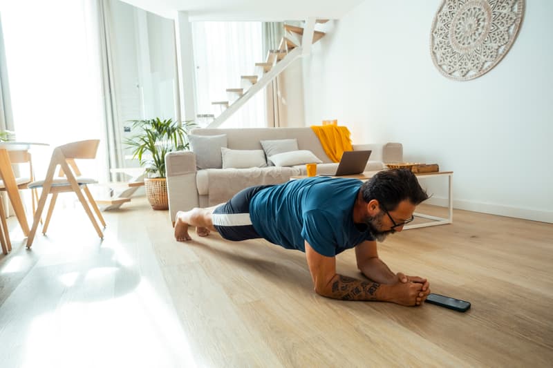 Mann macht im Wohnzimmer eine Unterarmstütze, vor ihm liegt sein Smartphone auf dem Boden