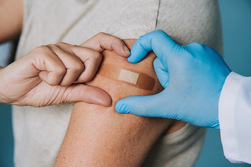Hand im blauen Handschuh drückt nach einer Impfung ein Pflaster auf den Oberarm einer Patientin