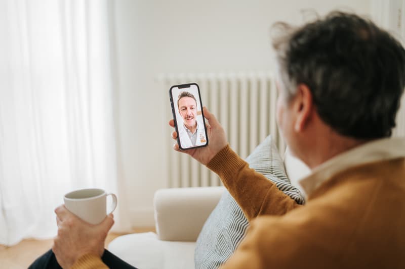 Mann mit Tasse auf dem Sofa spricht per Videoanruf auf dem Smartphone mit einem Arzt